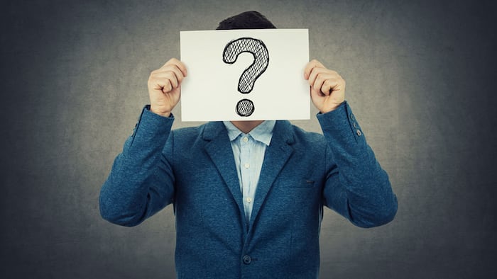 Businessman covering his face using a white paper with drawn question mark, like a mask, for hiding his identity. Isolated gray wall background.