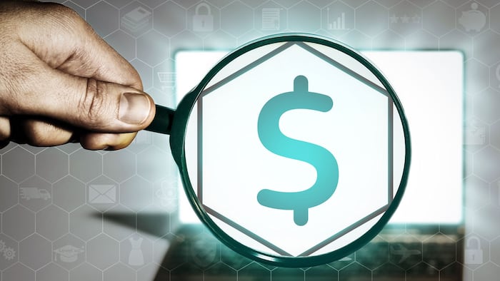 Hand holds the magnifying glass in front of an open notebook. Among the many icons, attention is focused on the money icon. Financial operations, banking, lending, etc.