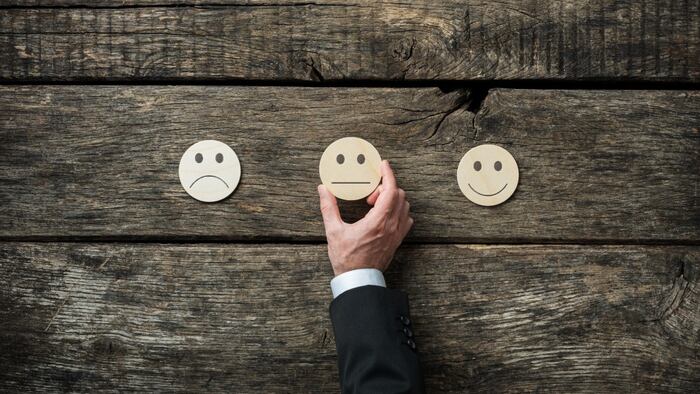 Wooden background with three drawn emoticon images in middle: sad, neutral and happy. Hand reaching out to place neutral emoticon.
