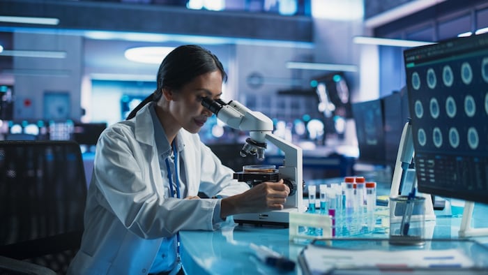Medicine Development Laboratory: Asian Female Scientist Using Microscope, Analyzes Petri Dish Sample. Big Pharmaceutical Lab with Specialists Conducting Biotechnology Research, Developing New Drugs.