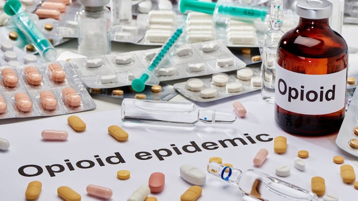 An ampoule Opioid next to it is a note written in English Opioid epidemic. All around, many tablets and syringes are scattered.