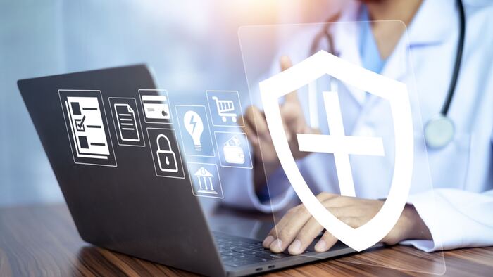 A doctor sits in front of a laptop, and various digital icons float above the screen, including a shield.